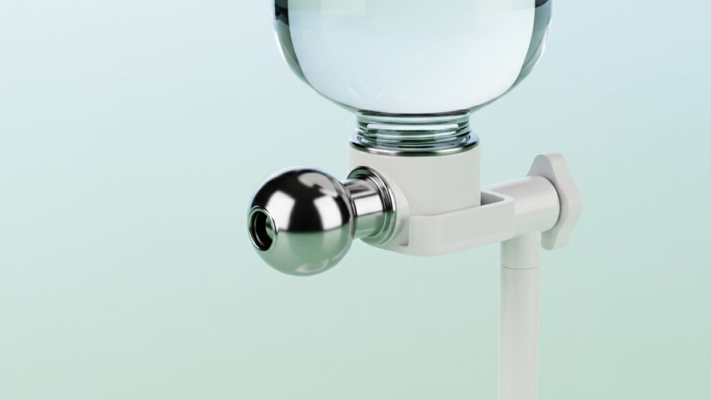 Close focado em um bico de bebedouro automático para animais de estimação. O dispositivo possui uma base de plástico branco que se prende a um suporte vertical. Um gargalo de vidro transparente e arredondado, cheio de água, está rosqueado na base. Do centro da base sai um cano de metal prateado e polido com uma ponta esférica e uma pequena abertura circular, projetado para lamber e evitar molhar o bigode de Shih Tzu. O fundo é um gradiente suave de azul-claro a verde-menta.